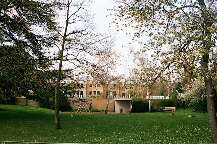 A large grassy area with scattered trees, a swing set, a wooden bench and a tan building in the background.