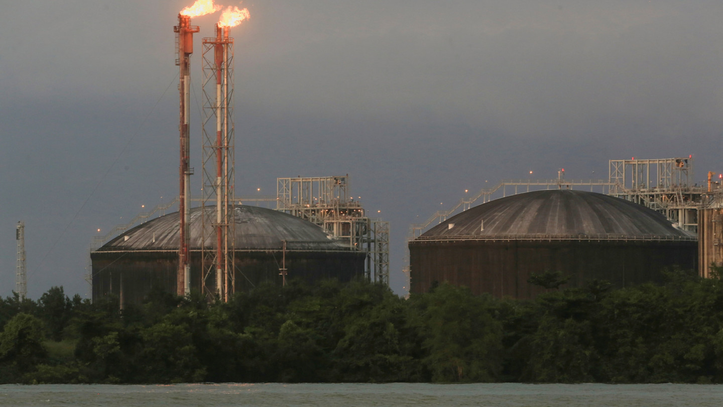Large storage tanks and tall gas flares emitting flames at the Atlantic LNG facility, with trees and water in the foreground.
