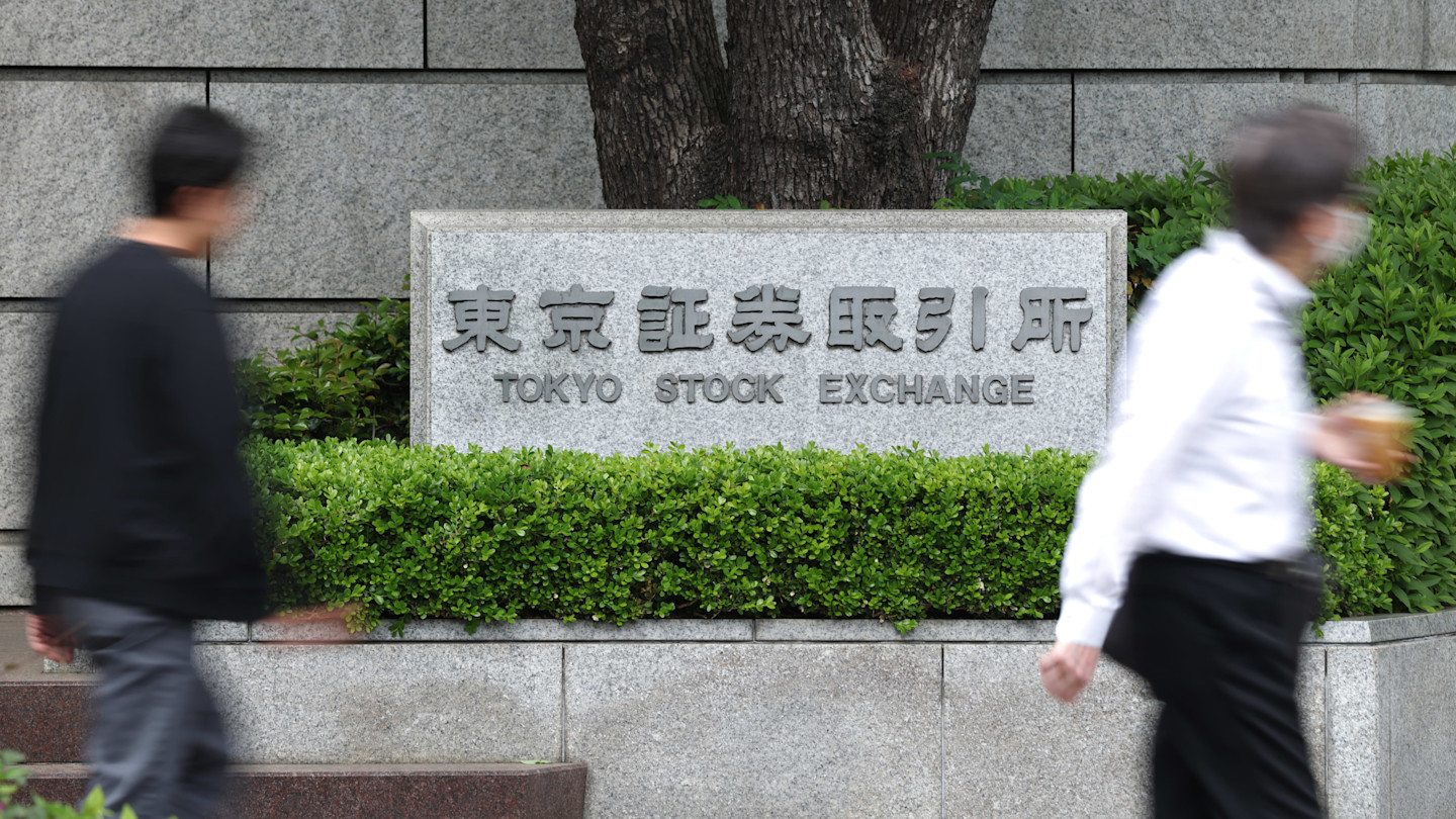 Sign for the Tokyo Stock Exchange in Japanese and English, with two blurred people walking past in the foreground.