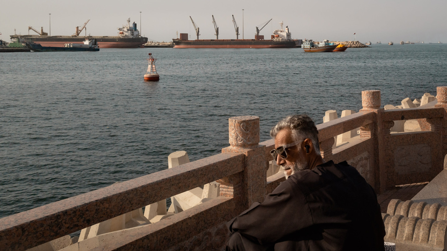 A man sits on a stone railing overlooking anchored oil tankers, cargo ships and fishing boats off the coast of Muscat, Oman