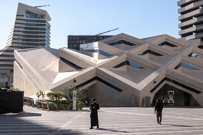 KAFD Grand Mosque with geometric modern architecture, two people walking in the plaza in front, surrounded by contemporary buildings.