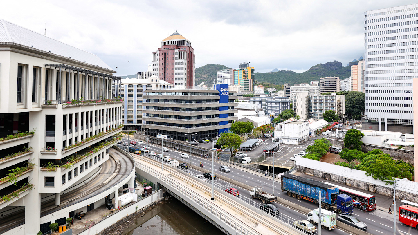 A view of Port Louis cityscape showing the Caudan Waterfront, office buildings, busy roads, and mountains in the background.