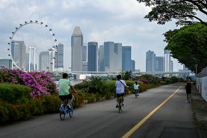 Several people cycle along a path lined with flowers and trees, with the Singapore city skyline and Singapore Flyer observation wheel in the background.