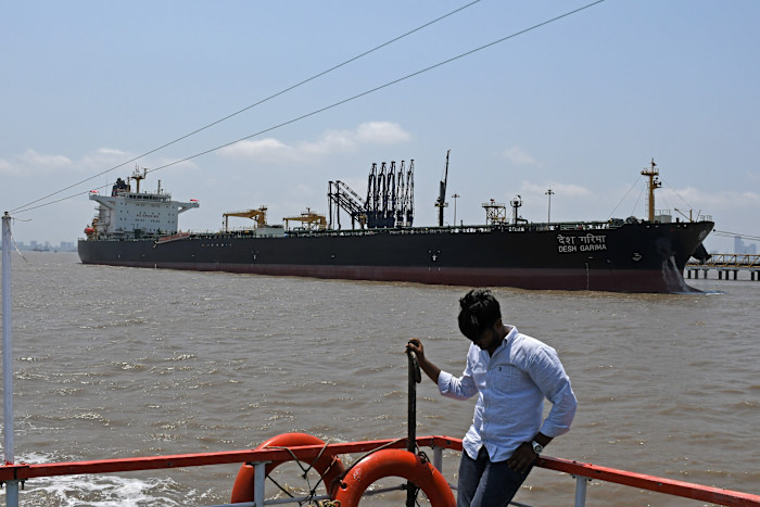 The crude oil tanker 'Desh Garima' is anchored at Mumbai port, with a person standing on a nearby vessel in the foreground.