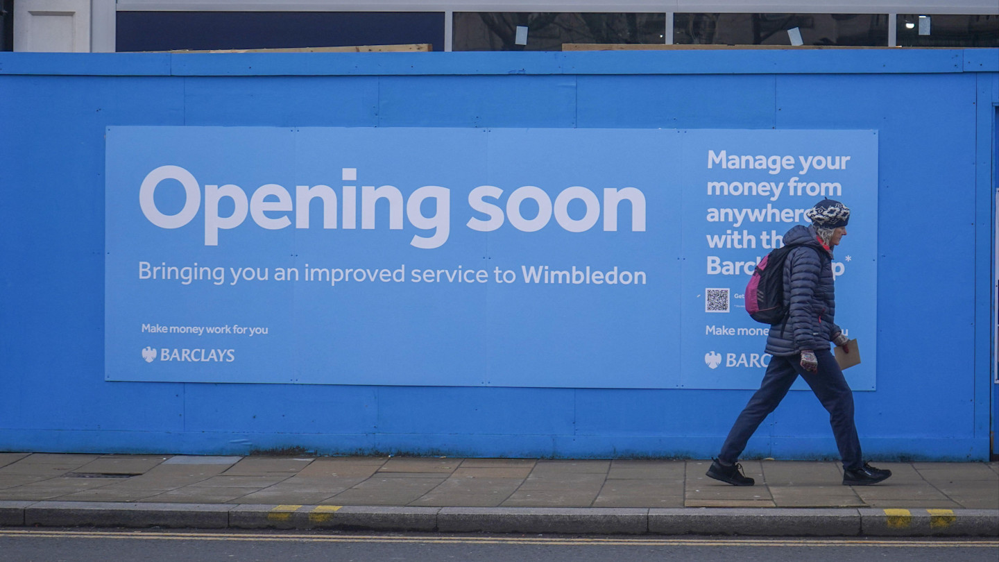 A person walks past a blue Barclays Bank hoarding announcing "Opening soon" for a new Wimbledon branch.