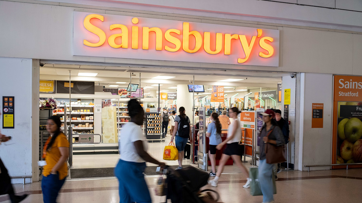 The entrance to Sainsbury's supermarket at Stratford Centre with people walking by and a large illuminated sign above the doors.