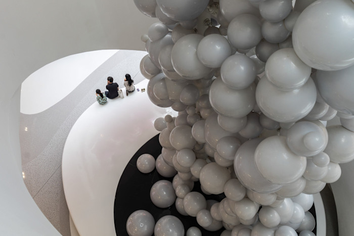 Three people sit on a white curved bench near a large sculptural installation made of clustered grey spheres.