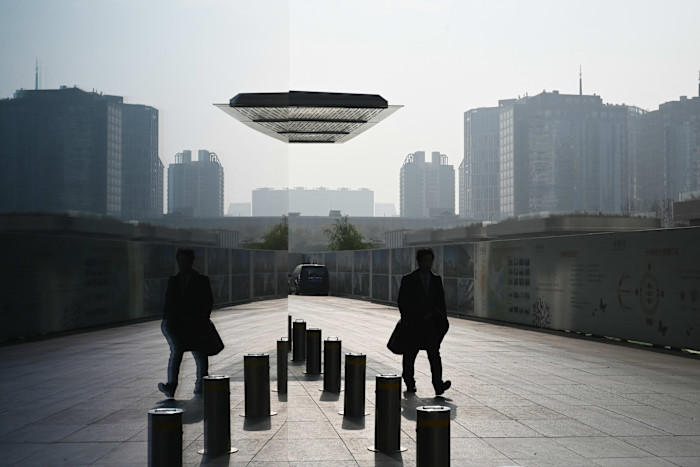 A man in a coat walks past metal bollards outside a building, with his reflection visible on a mirrored surface. Tall buildings are seen in the background.