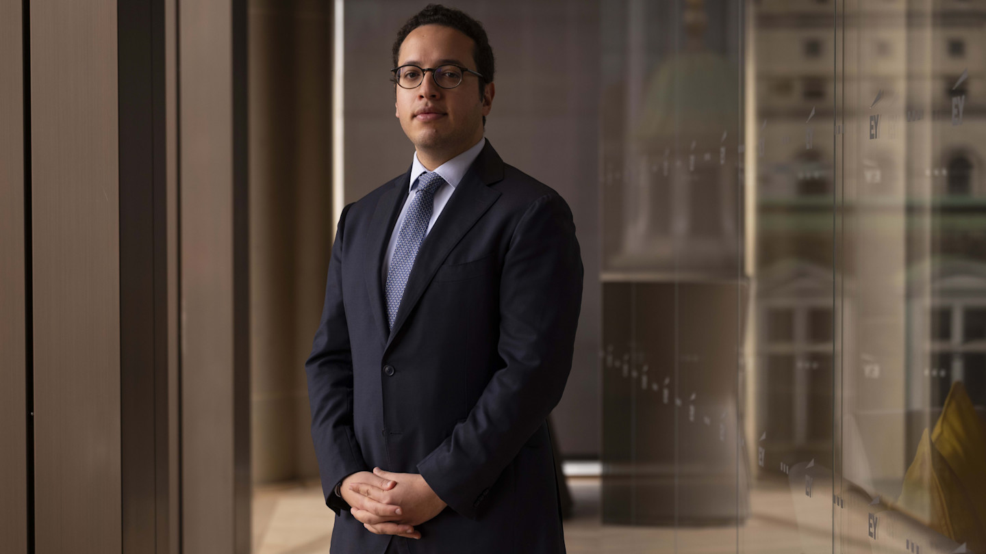 Hamza Lemssouguer stands indoors wearing a dark suit and tie, hands clasped, ahead of a television interview.