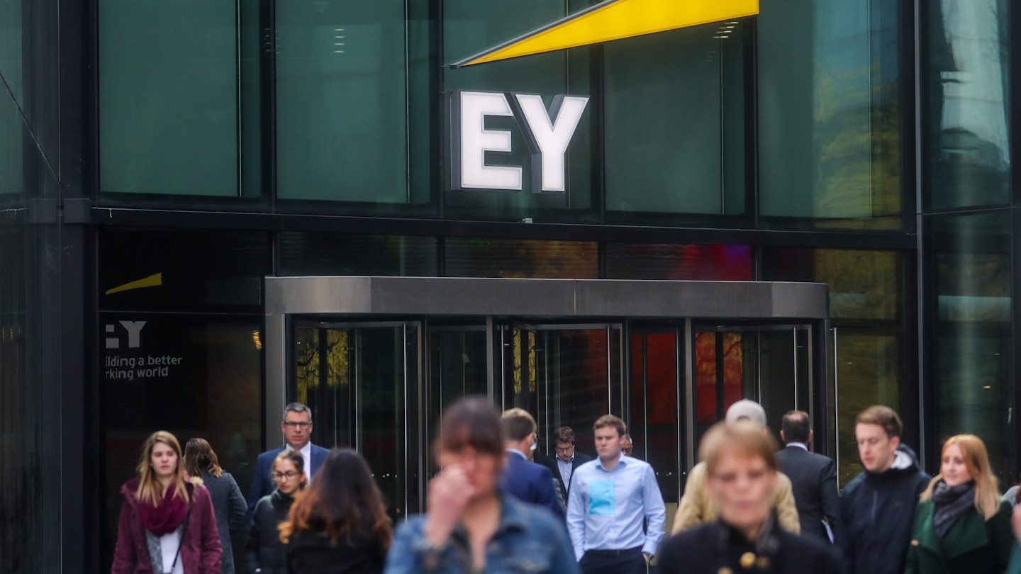 Pedestrians walk past the entrance to the Ernst & Young office in London, with a large EY sign above the doors.