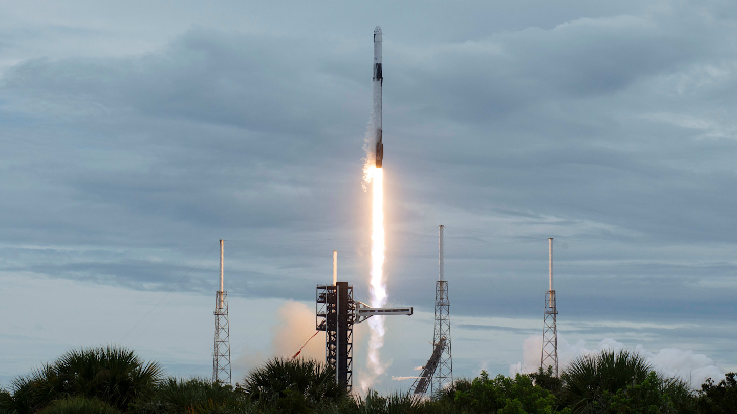 A SpaceX Falcon 9 rocket launches with bright flames and smoke, rising above the launchpad under a cloudy sky.