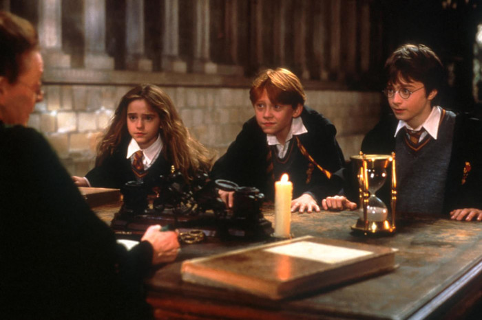 Emma Watson, Rupert Grint and Daniel Radcliffe as Hermione, Ron and Harry sit at a table listening to a teacher in a Hogwarts classroom.
