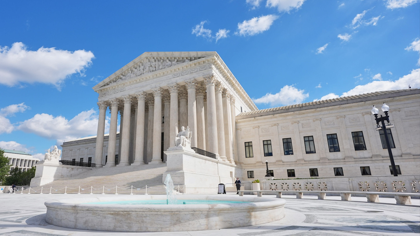 The Supreme Court building with its columned facade and a marble fountain in front under a blue sky with scattered clouds.