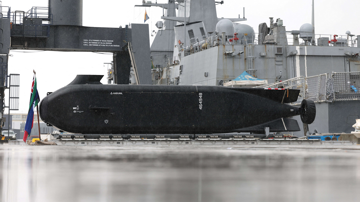 Anduril Ghost Shark extra-large autonomous undersea vehicle displayed on a naval base pier, with a large naval ship in the background.