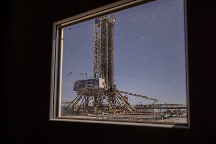 A drilling rig stands at the Vista Energy Bajada del Palo Oil and Gas Field, seen through a window under a clear sky.