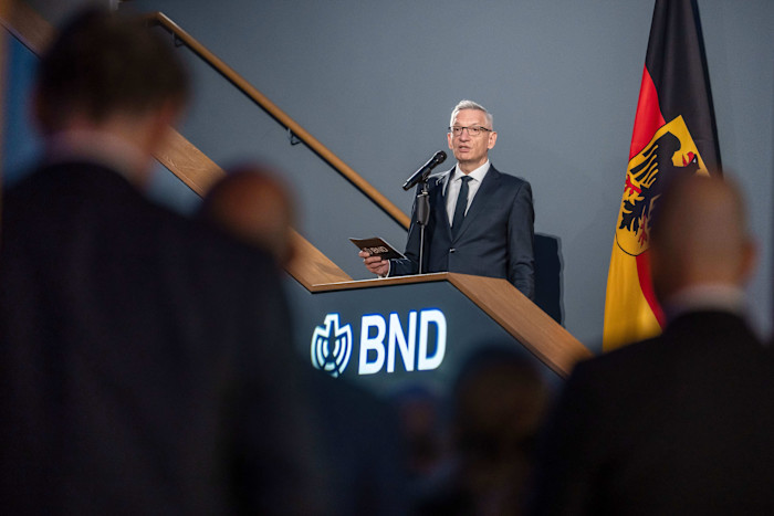 Martin Jäger stands at a podium marked "BND," speaking at a microphone with the German flag behind him.