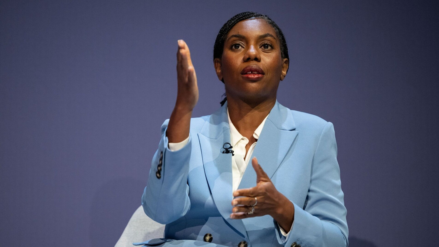 Kemi Badenoch gestures with one hand raised while speaking, wearing a light-blue suit against a plain background.