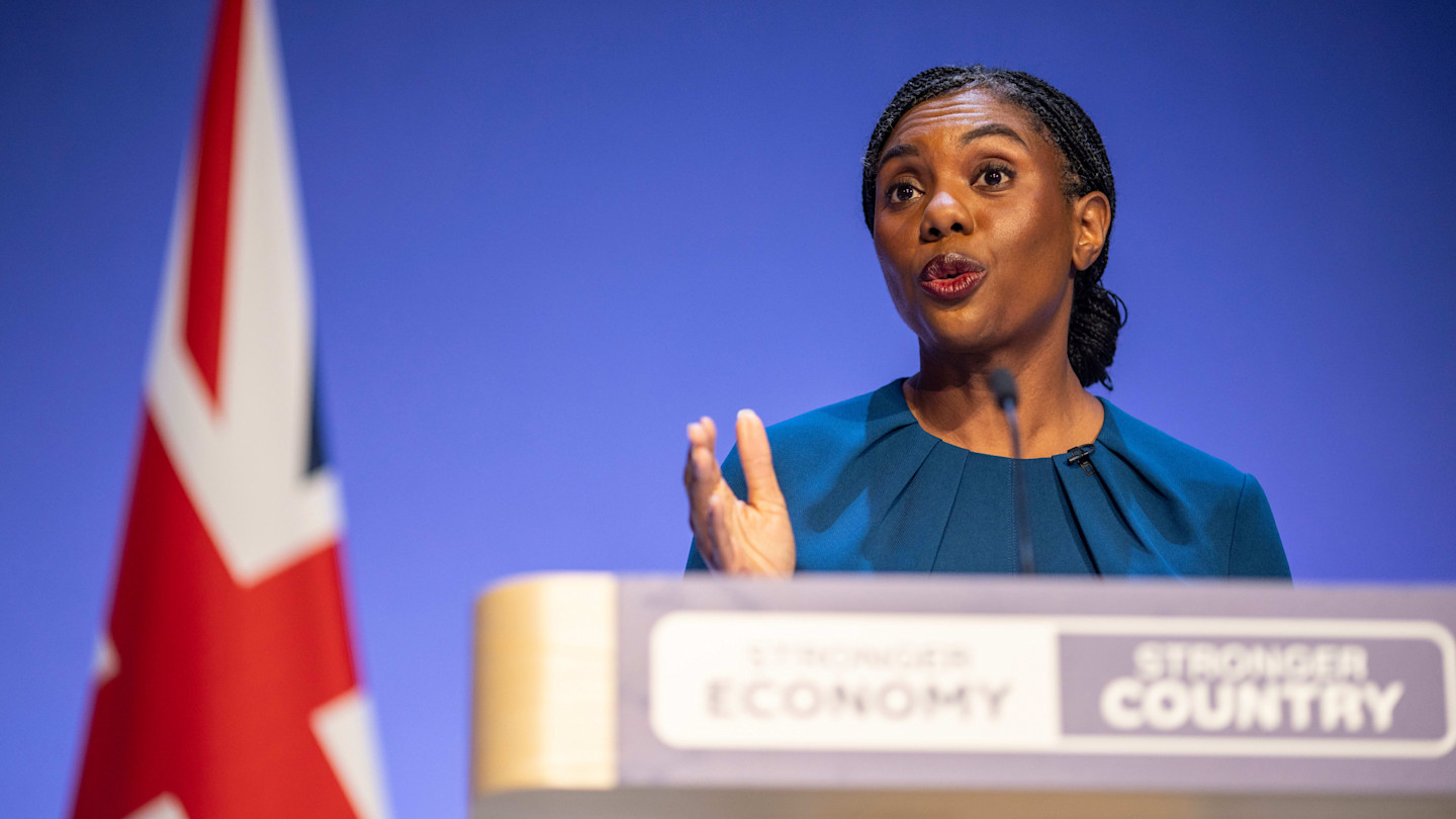 Kemi Badenoch speaks at a podium with "Stronger Economy" and "Stronger Country" signs, a UK flag visible beside her.