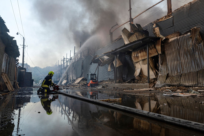 A firefighter sprays water on a heavily damaged warehouse with collapsed metal walls and rising smoke after a missile attack in Kyiv.
