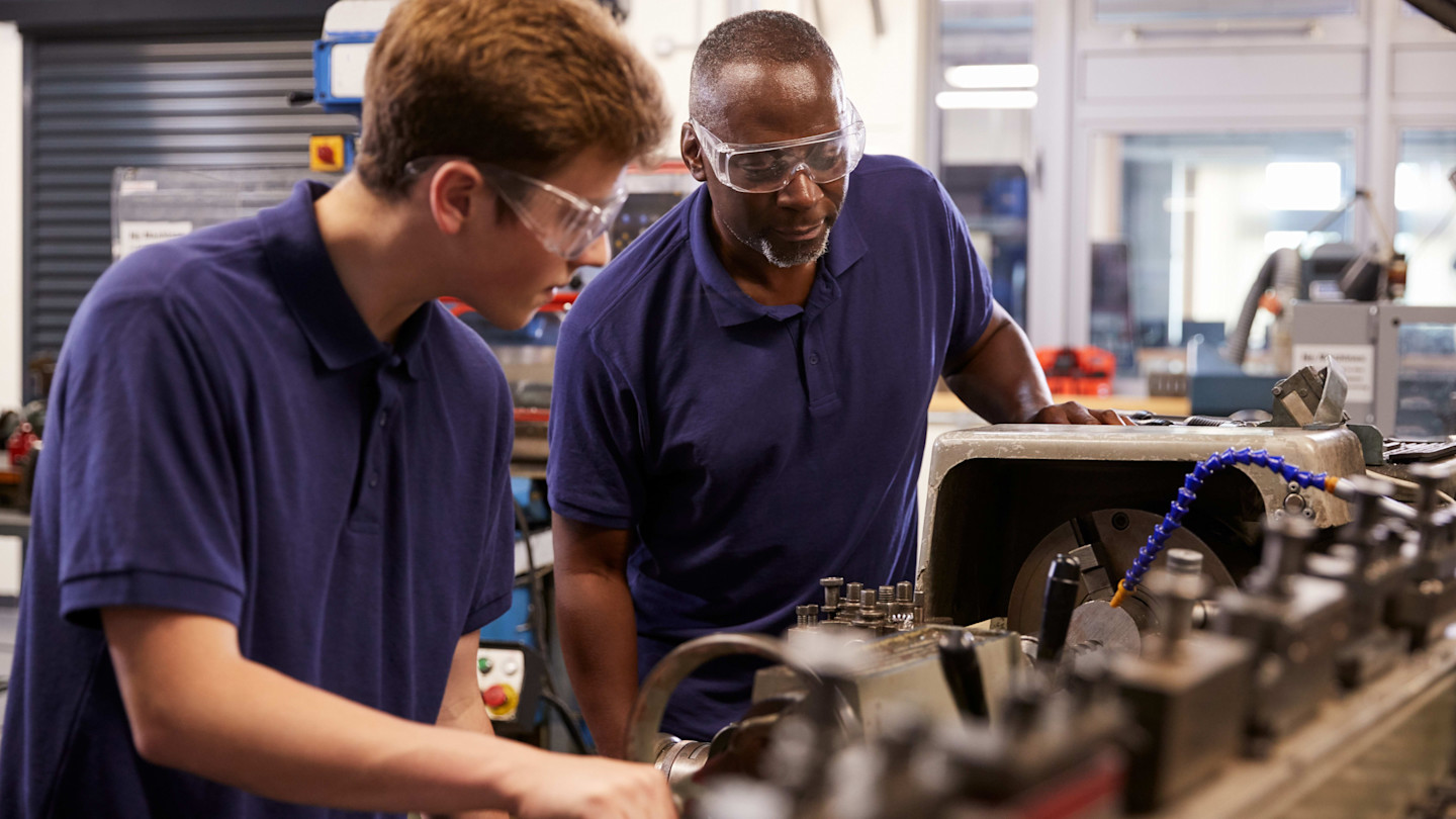 An engineer supervises a teenage apprentice operating a lathe machine. Both wear safety goggles and blue shirts in a workshop.