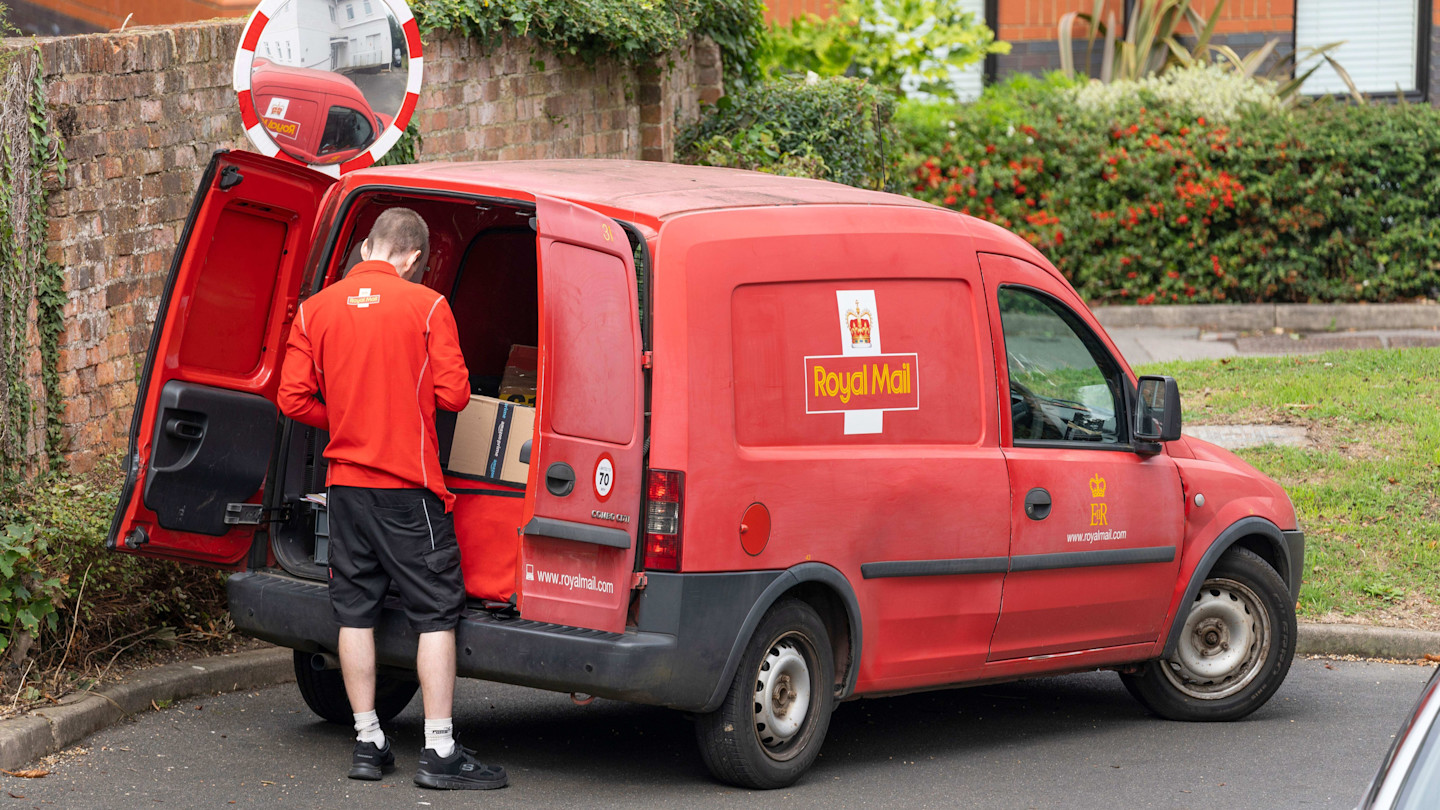 A Royal Mail delivery worker stands at the open back of a red Royal Mail van, organizing parcels for delivery.