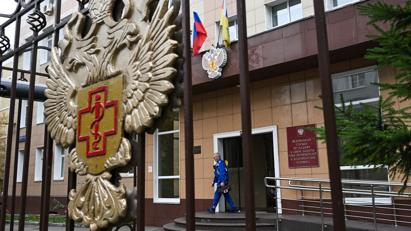 A maintenance worker in blue uniform exits the Rospotrebnadzor headquarters in Moscow, seen through a gate with the agency's emblem.