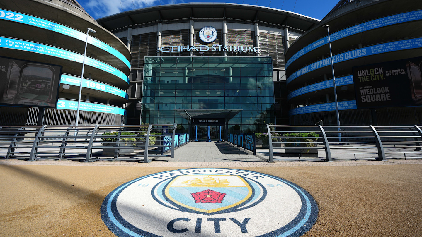 The main entrance of the Etihad Stadium, with the Manchester City crest on the pavement in the foreground.