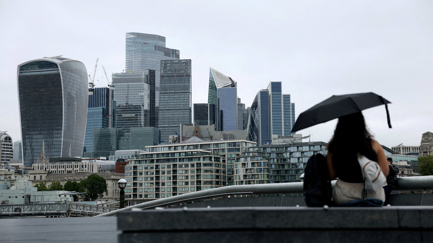 A woman with an umbrella sits facing the City of London skyline on a cloudy day.