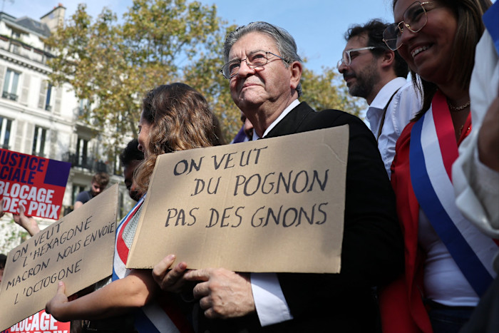 Jean-Luc Mélenchon holds a cardboard sign reading "On veut du pognon pas des gnons" during a rally, surrounded by supporters.