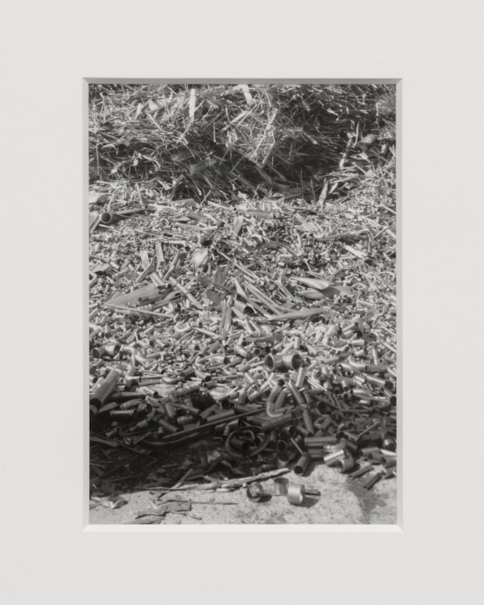 A black and white photograph by Nat Faulkner showing a dense pile of metal pipes and fittings scattered on the ground.
