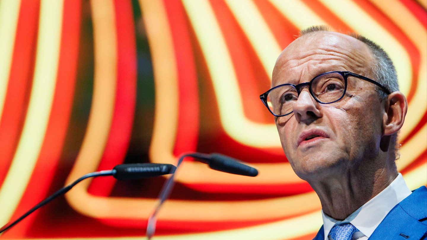 Friedrich Merz speaks at a podium during the Germany Summit, with colourful abstract lines in the background.