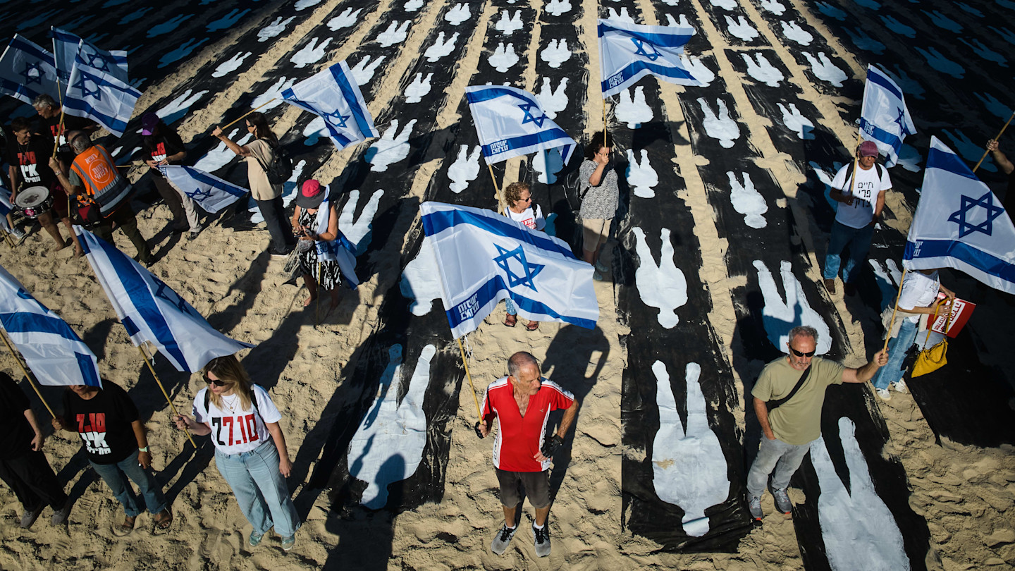 People holding Israeli flags stand on a beach next to an installation of white human silhouettes marking the third anniversary of the Oct 7 2023 Hamas attack.