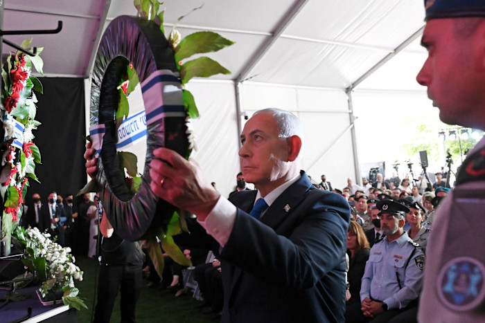 Benjamin Netanyahu solemnly places a wreath at a memorial ceremony, with uniformed officials and attendees seated behind him.