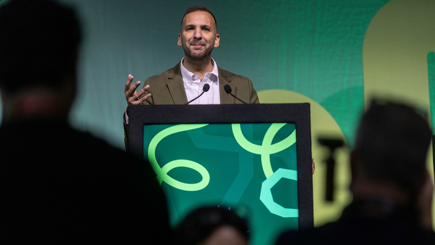Zack Polanski speaks at a podium with a green patterned backdrop at the Green Party conference.