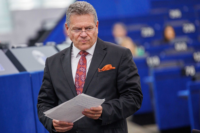 Maroš Šefčovič holds papers and speaks during a European Parliament session. Blue seats are visible in the background.