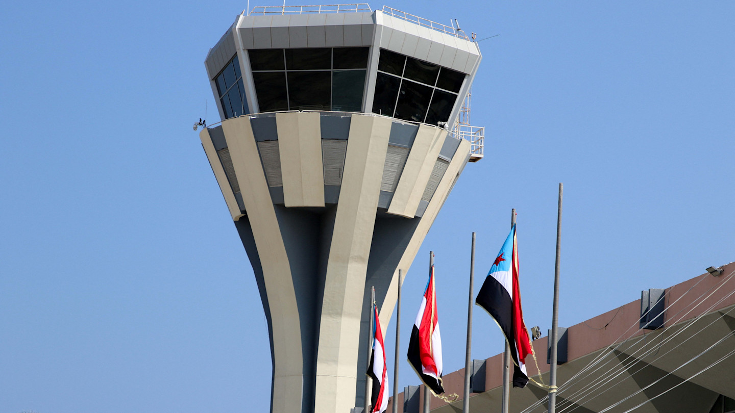 The control tower at Aden Airport with four flags in front, set against a clear blue sky.