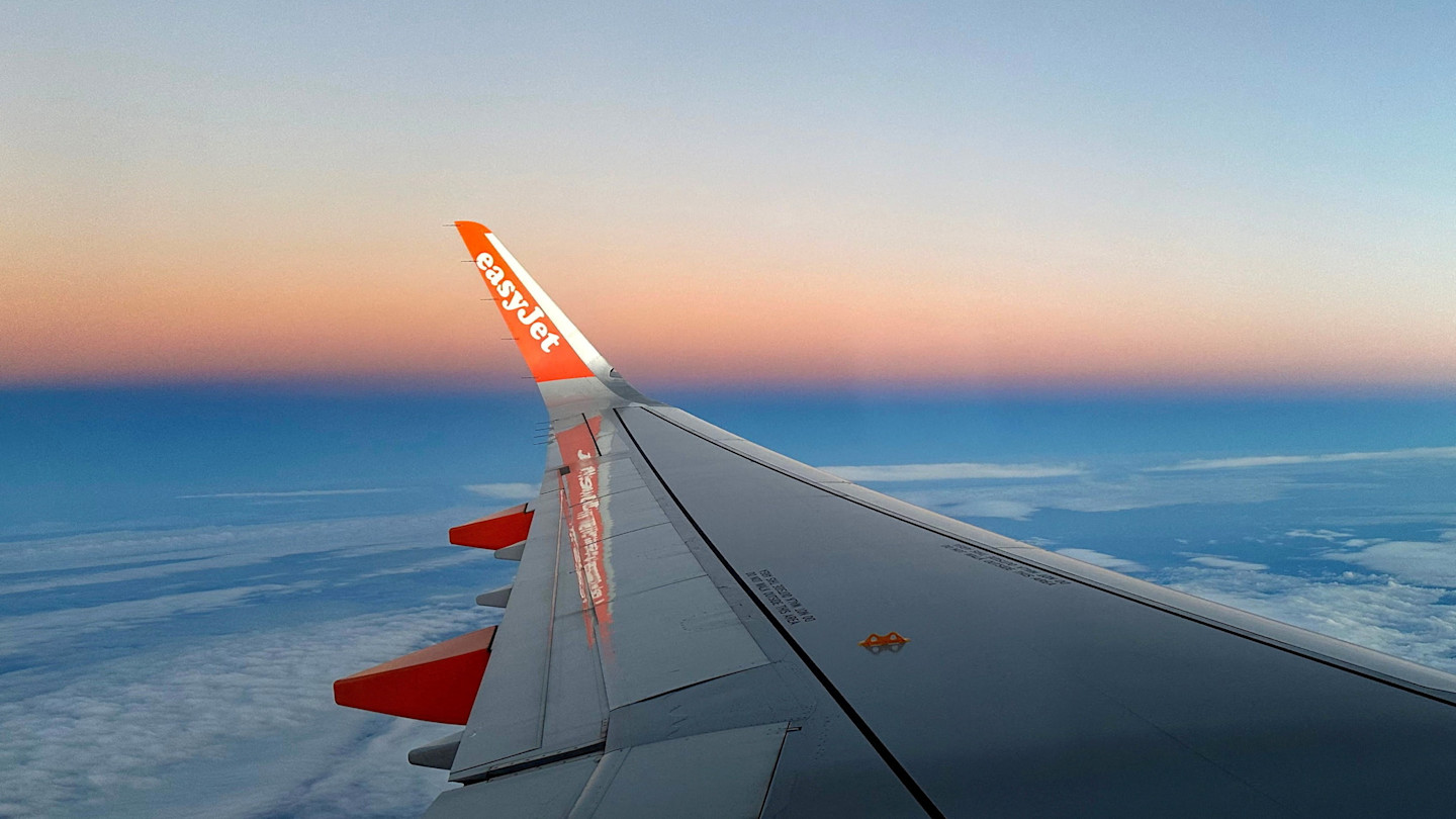 The wing of an EasyJet aeroplane is seen against a colourful sunset sky above the clouds.