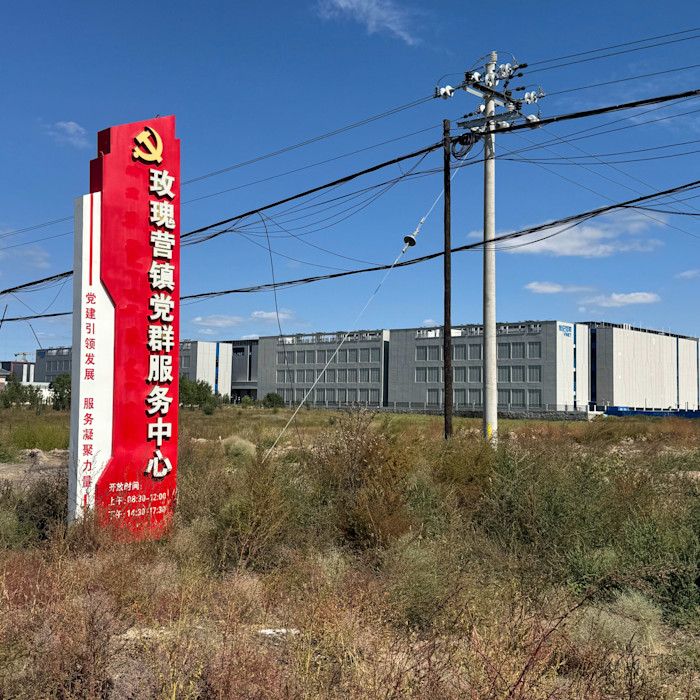 Large, grey data centre buildings with rooftop infrastructure stand behind overgrown grass and a red sign with Chinese text and a Communist Party emblem in Ulanqab, Inner Mongolia.