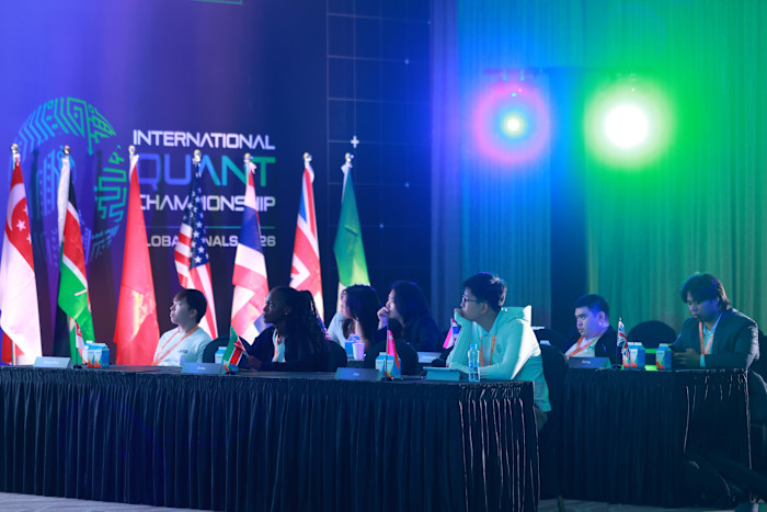 A group of participants sit at tables with small national flags in front of them during the International Quant Championship 2026, with international flags and colourful lights in the background.