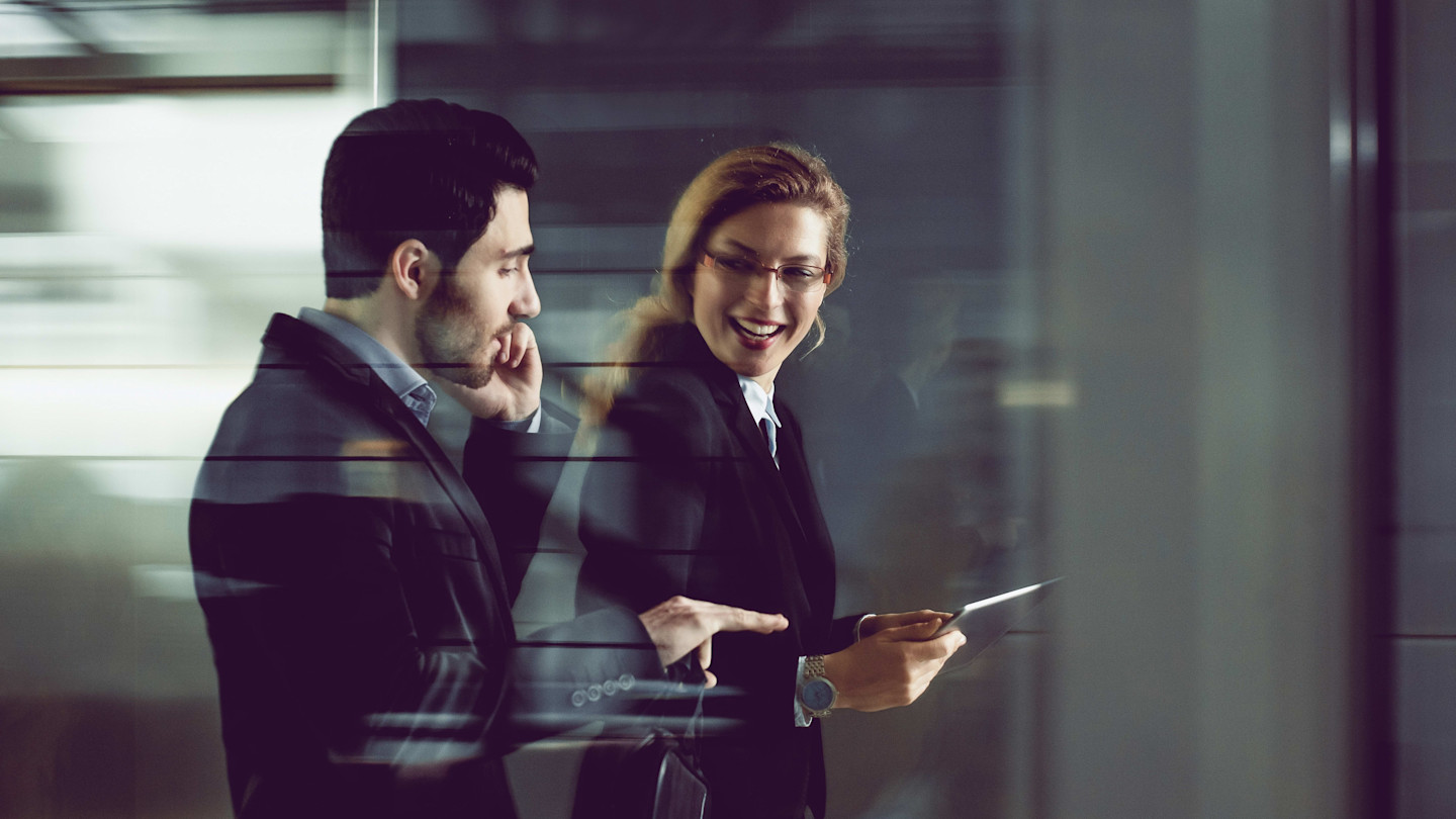 Two business professionals in suits walking together; one is talking on the phone, the other is smiling and holding a tablet.