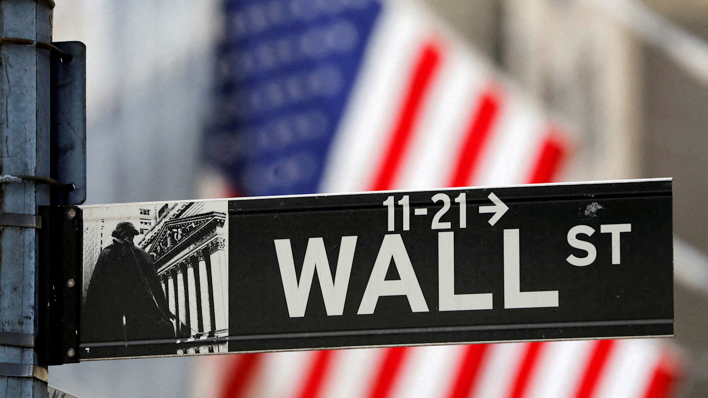 Wall Street street sign with part of the New York Stock Exchange building and a blurred American flag in the background.
