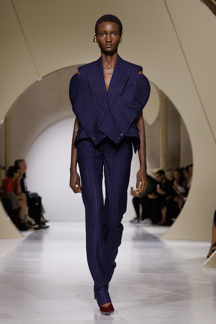 A model walks the runway in a navy pinstripe suit with a sculptural jacket featuring crossed sleeves and cutaway shoulders.