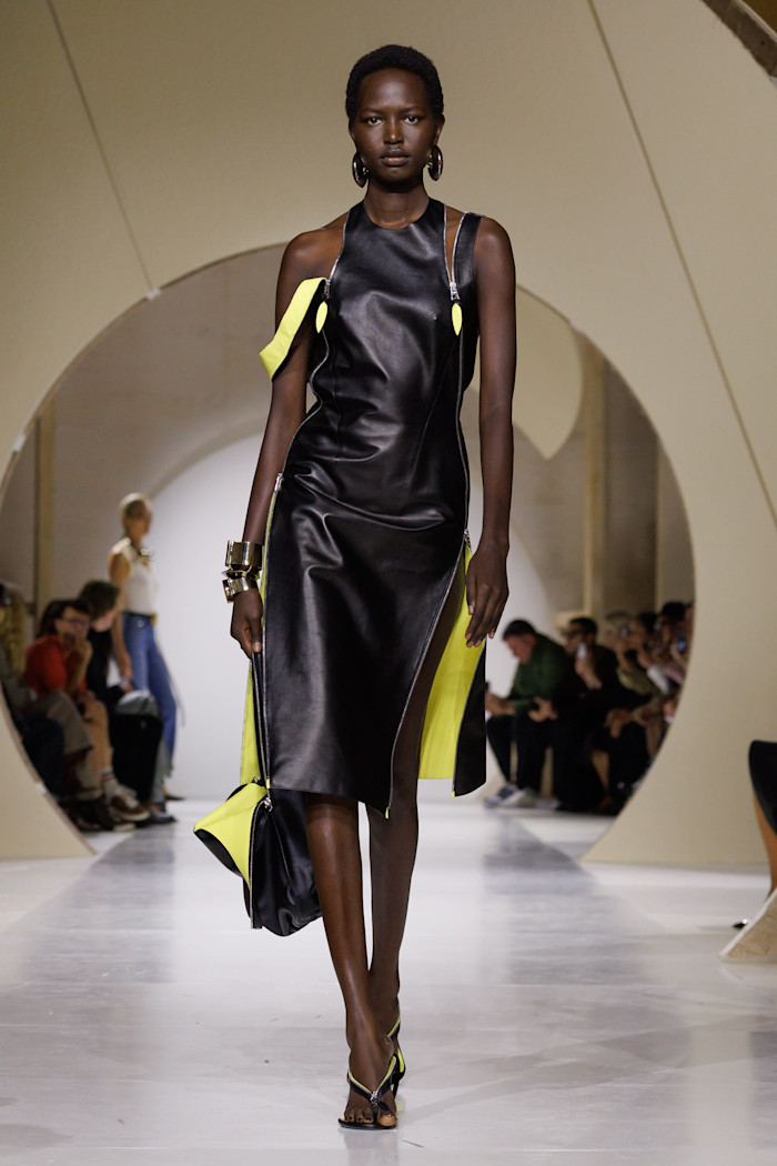 A model walks the runway in a sleeveless black leather midi dress with bright yellow panels, carrying a matching black and yellow bag.