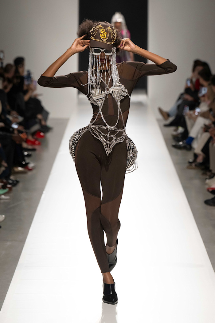 A model walks the runway in a sheer, form-fitting brown bodysuit adorned with metallic chains, a baseball cap, and sunglasses draped with beaded fringe.