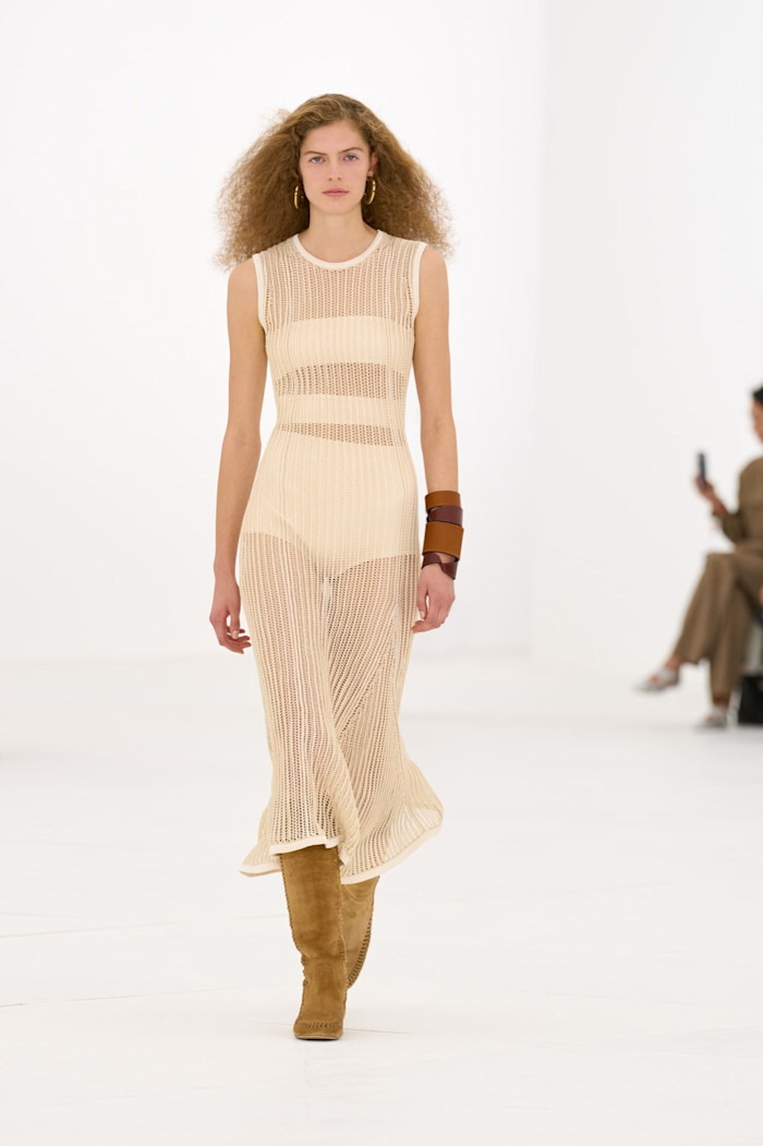 A model walks the runway in a sleeveless cream open-knit midi dress, paired with tan knee-high boots and wide leather cuffs.