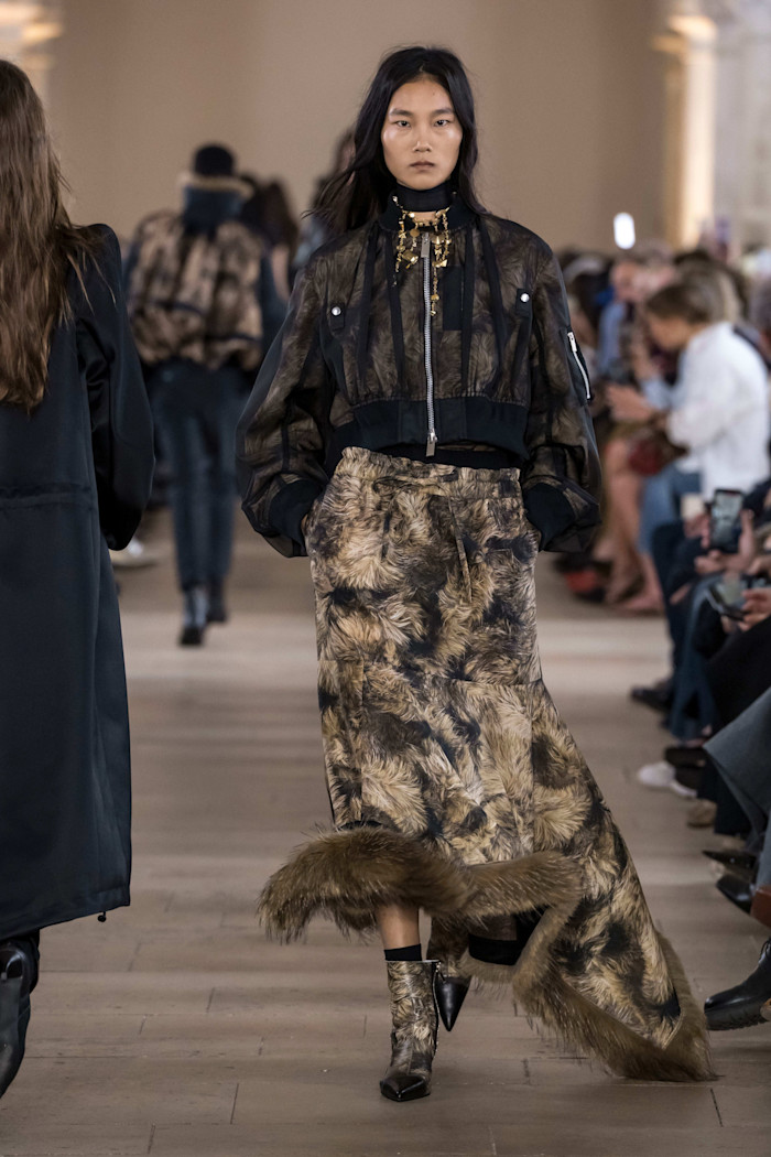 A model walks the runway in a sheer black cropped jacket and a long brown patterned skirt with faux-fur trim, paired with matching boots.