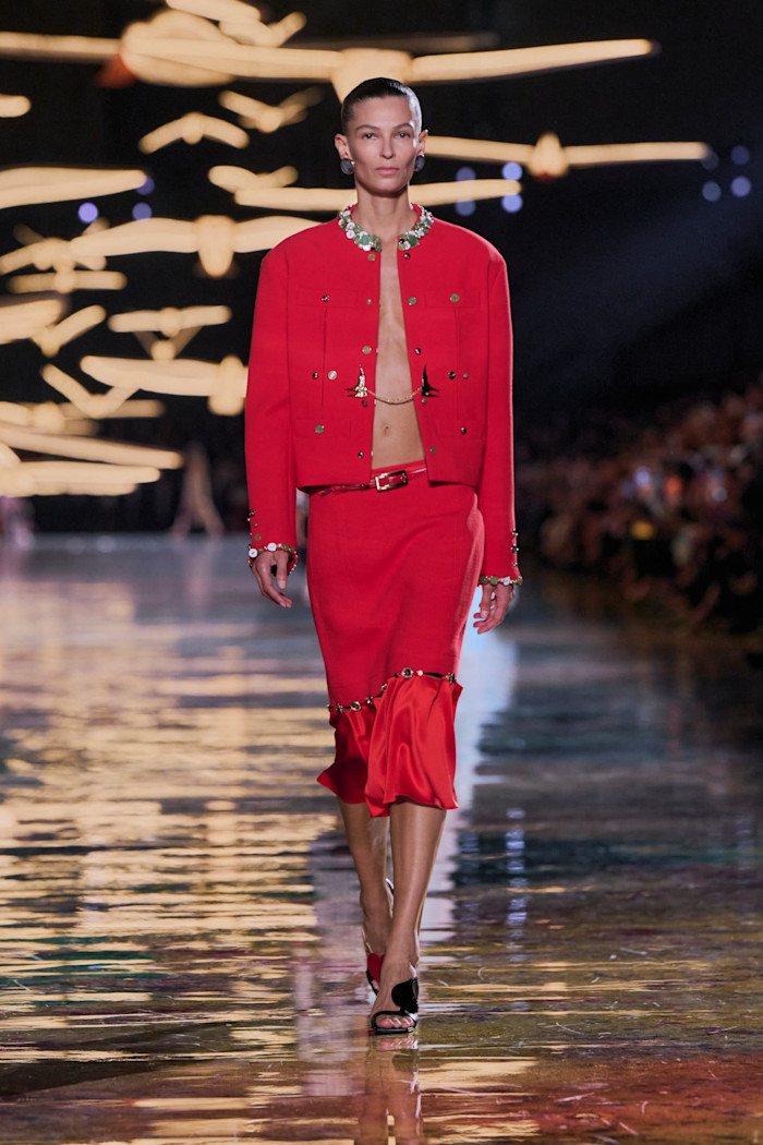 A model walks the runway in a cropped red jacket and matching knee-length skirt, embellished with beads and jewellery.
