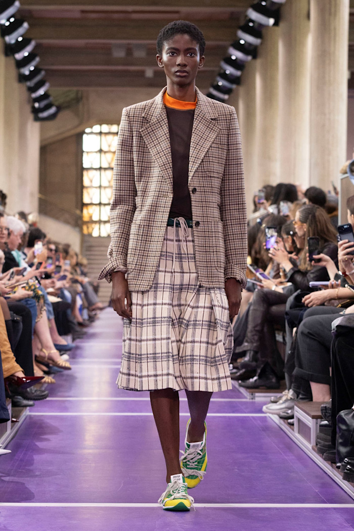 A model walks the runway in a checked blazer and matching pleated knee-length shorts, paired with a brown top and colourful trainers.