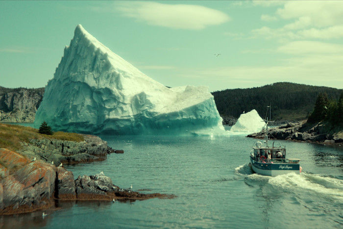 A fishing boat travels through icy waters near a massive iceberg, with rocky shores and forested hills in the background.