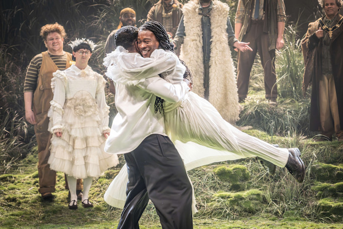 Two actors embrace joyfully center stage in period costumes, surrounded by cast members in whimsical attire against a woodland set.